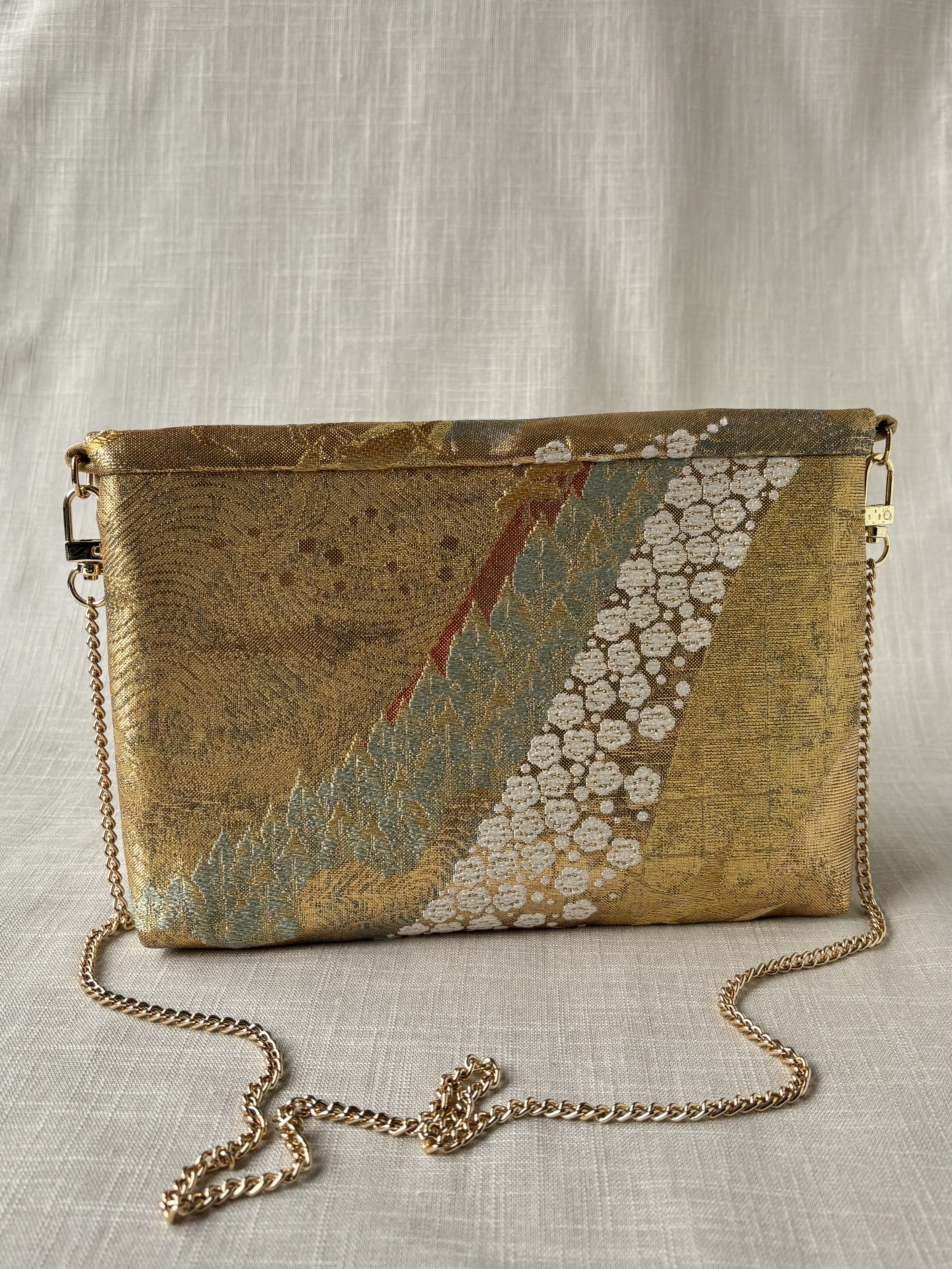 Back view of clutch with a pattern of pine and plum on a shimmering golden background, the clutch stand before cream coloured textured linen background.