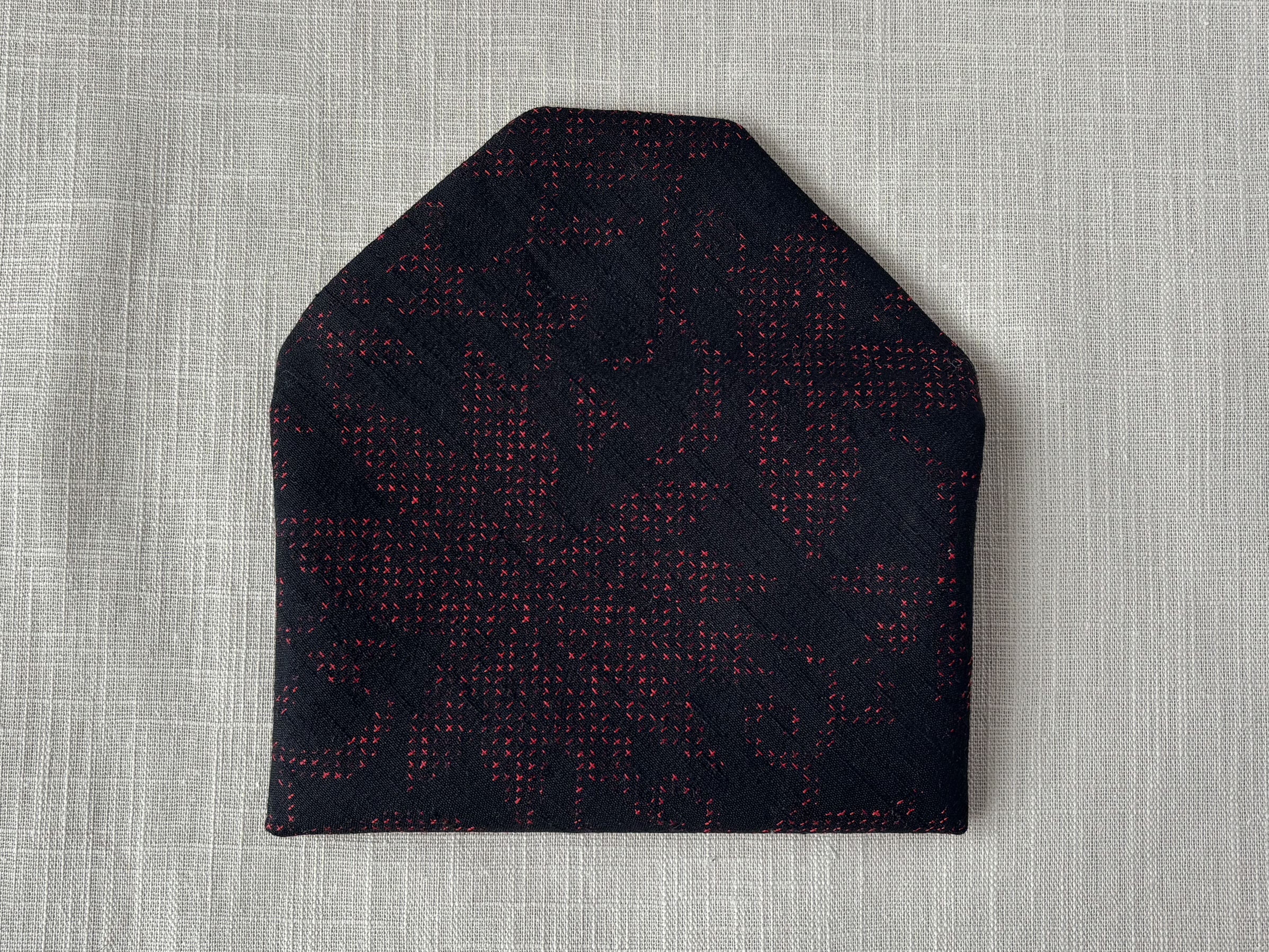 Back view of the matte black sukiya bag with crimson cross stitch pattern on a cream coloured textured linen background.